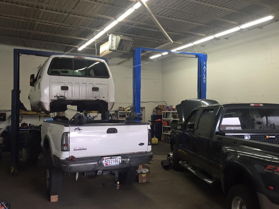 Two trucks in a garage, one on a lift. White truck bed is raised, black truck is beside it.