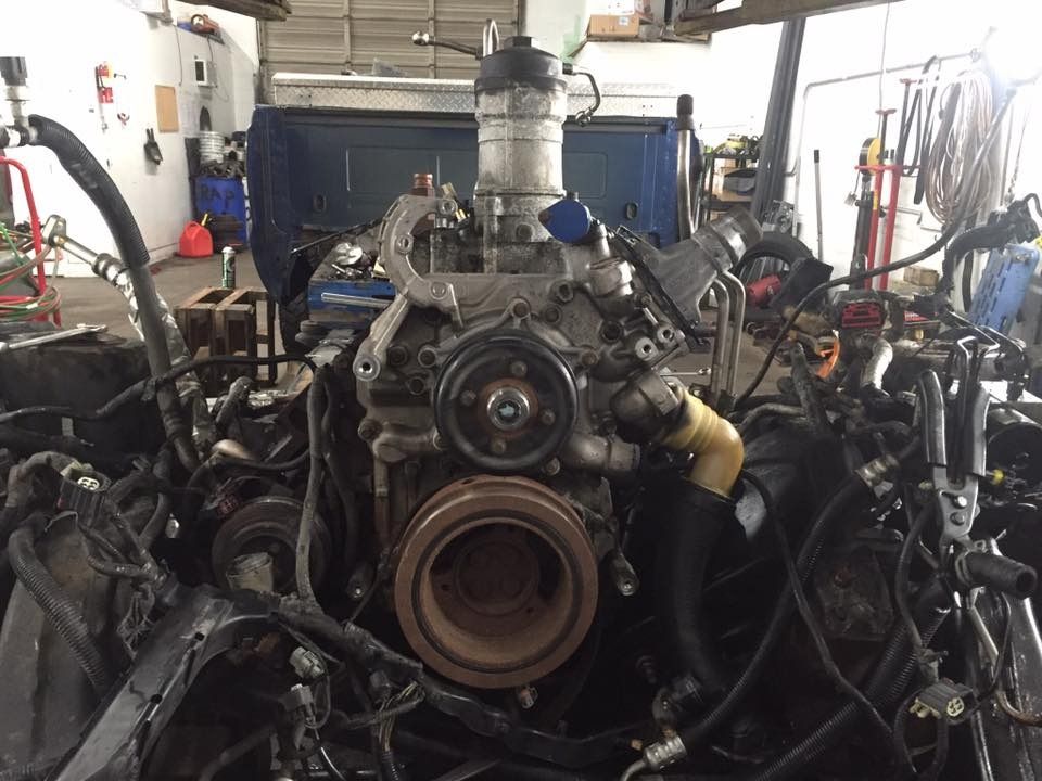 A car engine in a repair shop, various parts visible, black and gray with some yellow hoses.