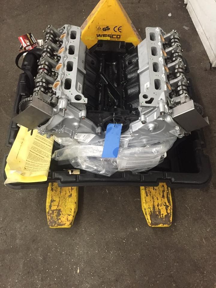 Engine parts on a pallet; metallic gray engine components, wrapped in plastic; yellow pallet jacks.