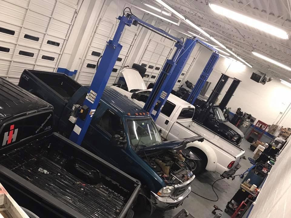 Trucks in a repair shop, one on a lift, hoods open. Blue, white, and black vehicles in a well-lit garage.