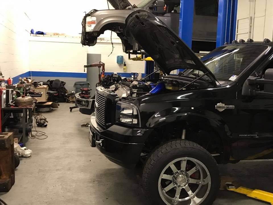 Black truck with open hood in a repair shop, another vehicle's front end suspended overhead.