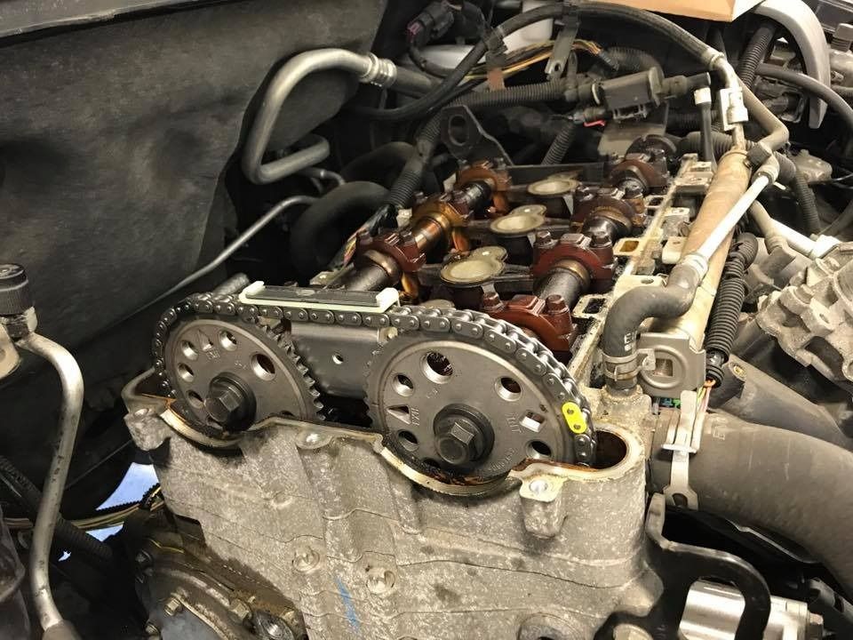 Engine with timing chain exposed, showing gears and camshafts, inside a vehicle's engine bay.