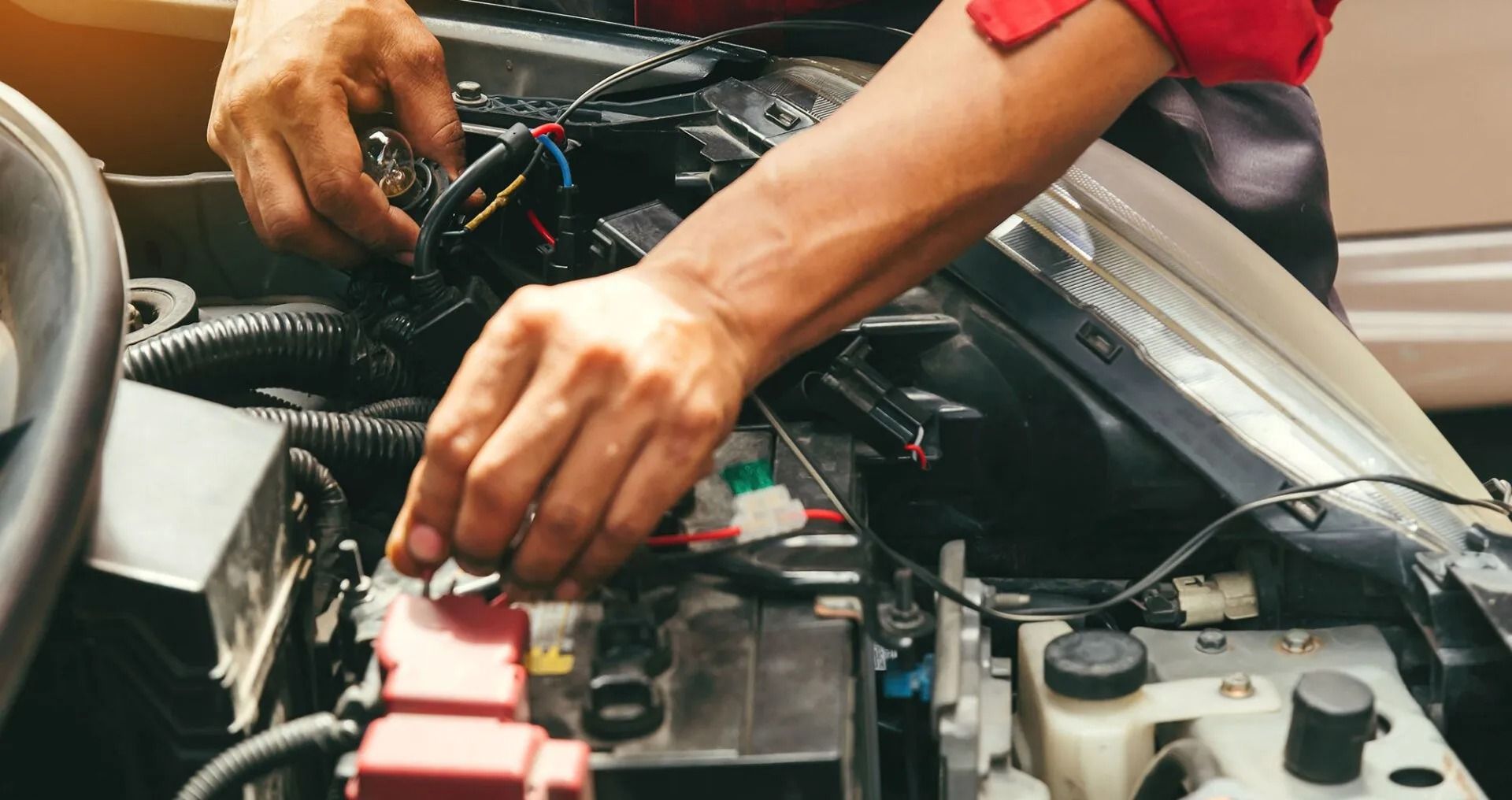Mechanic working on a car engine, connecting wires, in a well-lit outdoor setting.