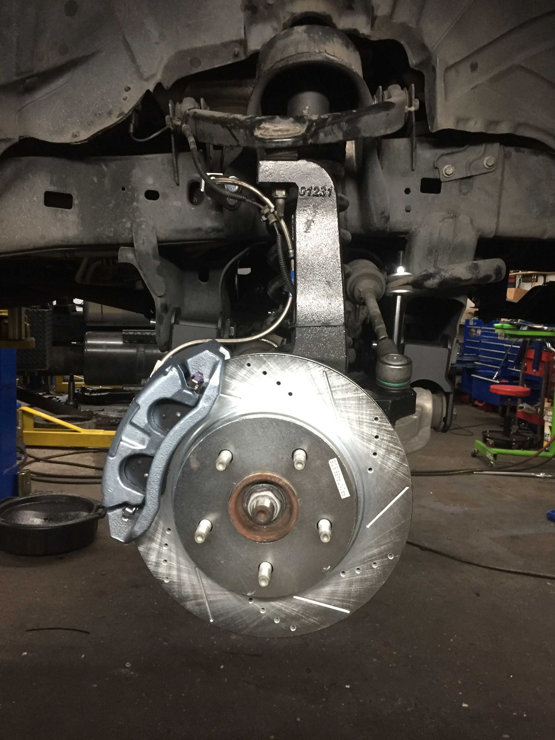 Car's front wheel assembly undergoing repair. Grey caliper, drilled and slotted rotor visible.