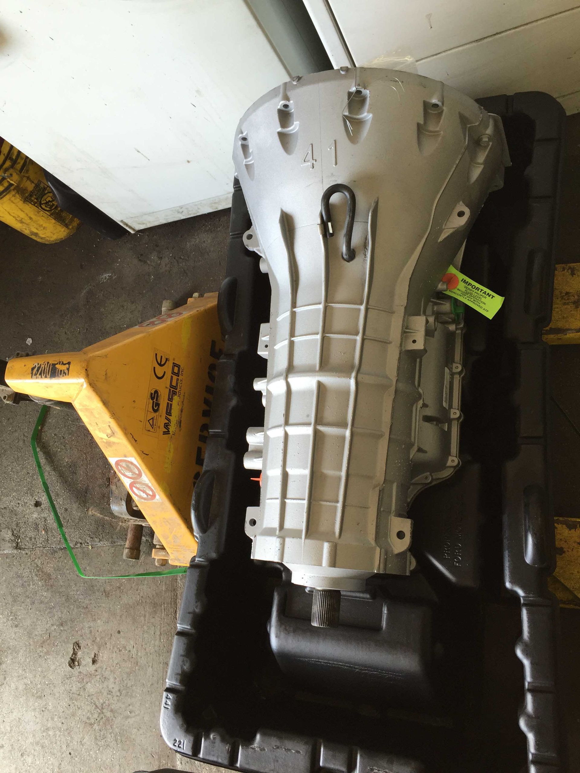 Silver transmission in a black plastic container on a pallet jack, against a white wall.