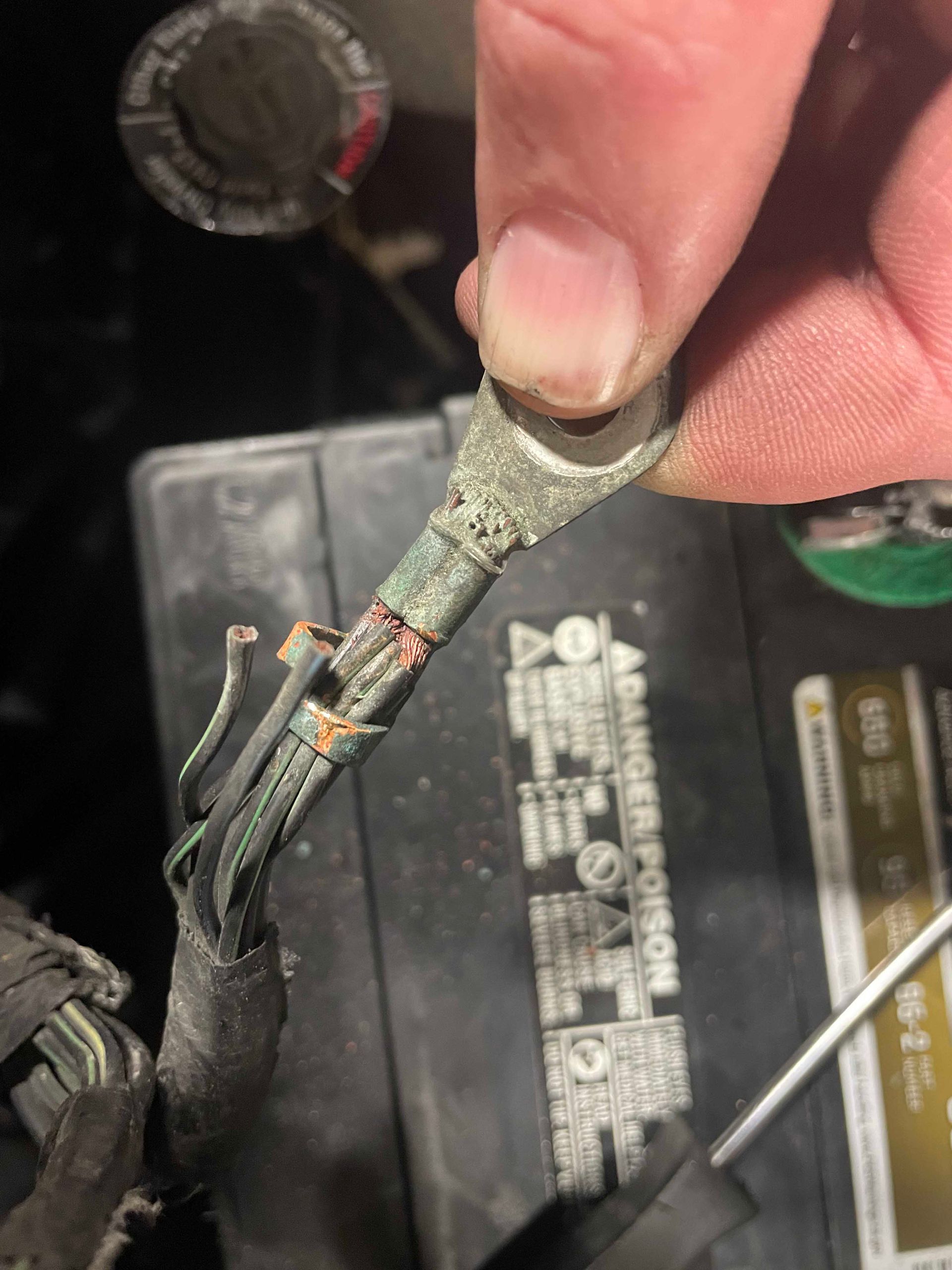 Corroded battery terminal wire held by a hand. Exposed wires, black electrical tape, and a battery in the background.