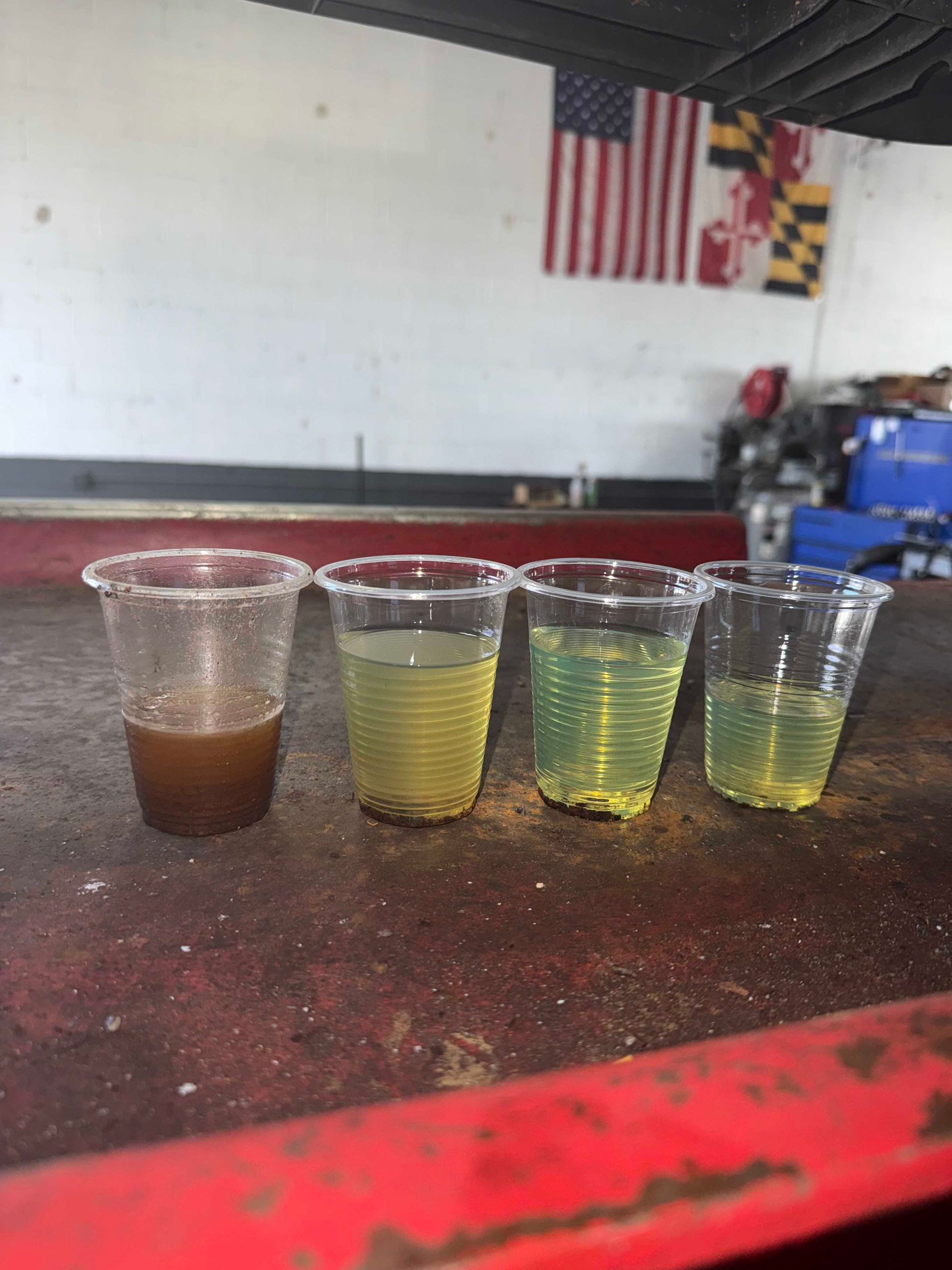 Four clear cups with varying shades of yellow liquid, placed on a red surface. The first cup is brown. An American flag hangs in the background.
