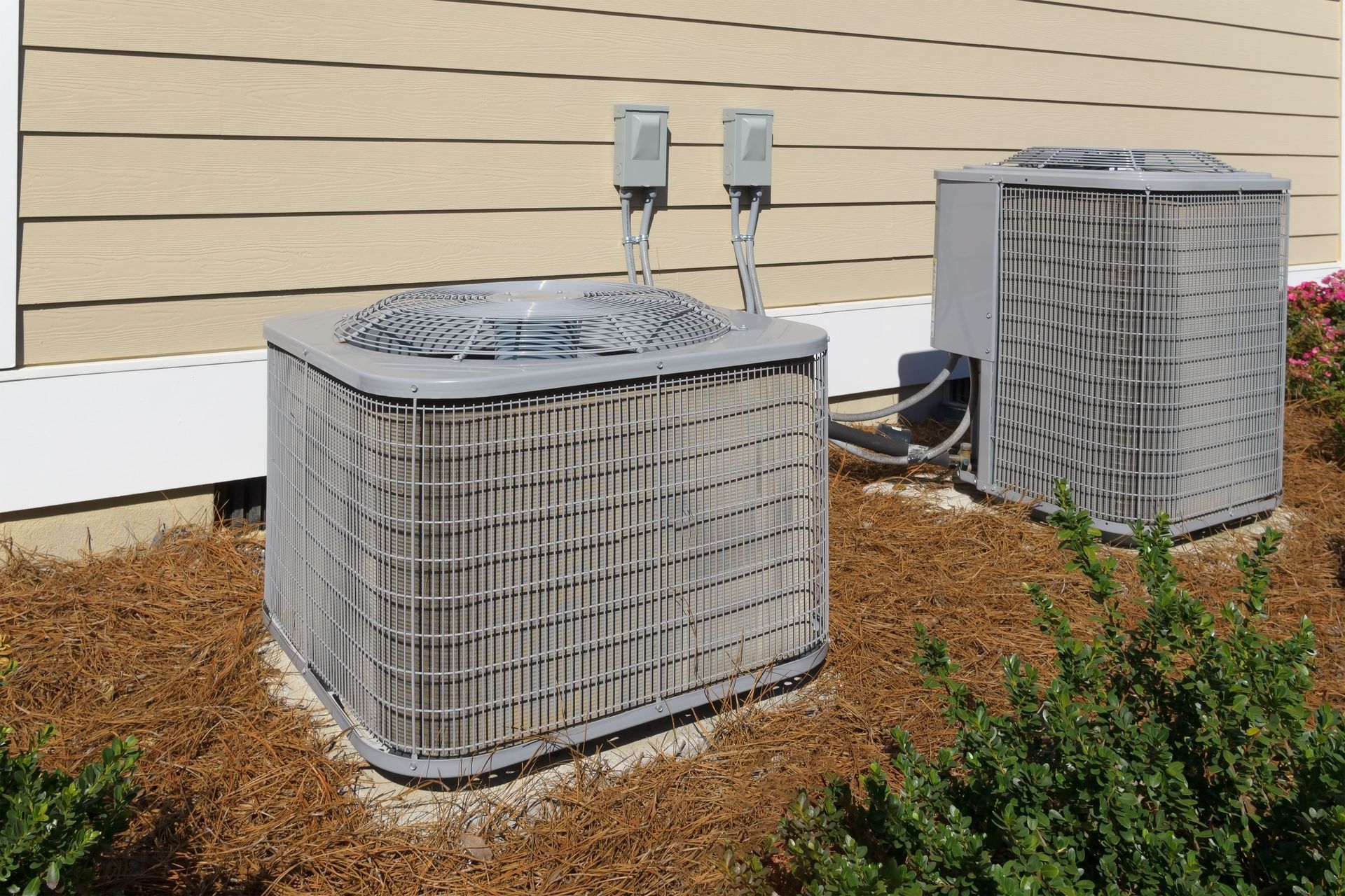 Two outdoor air conditioning units beside a beige house wall and brown mulch.