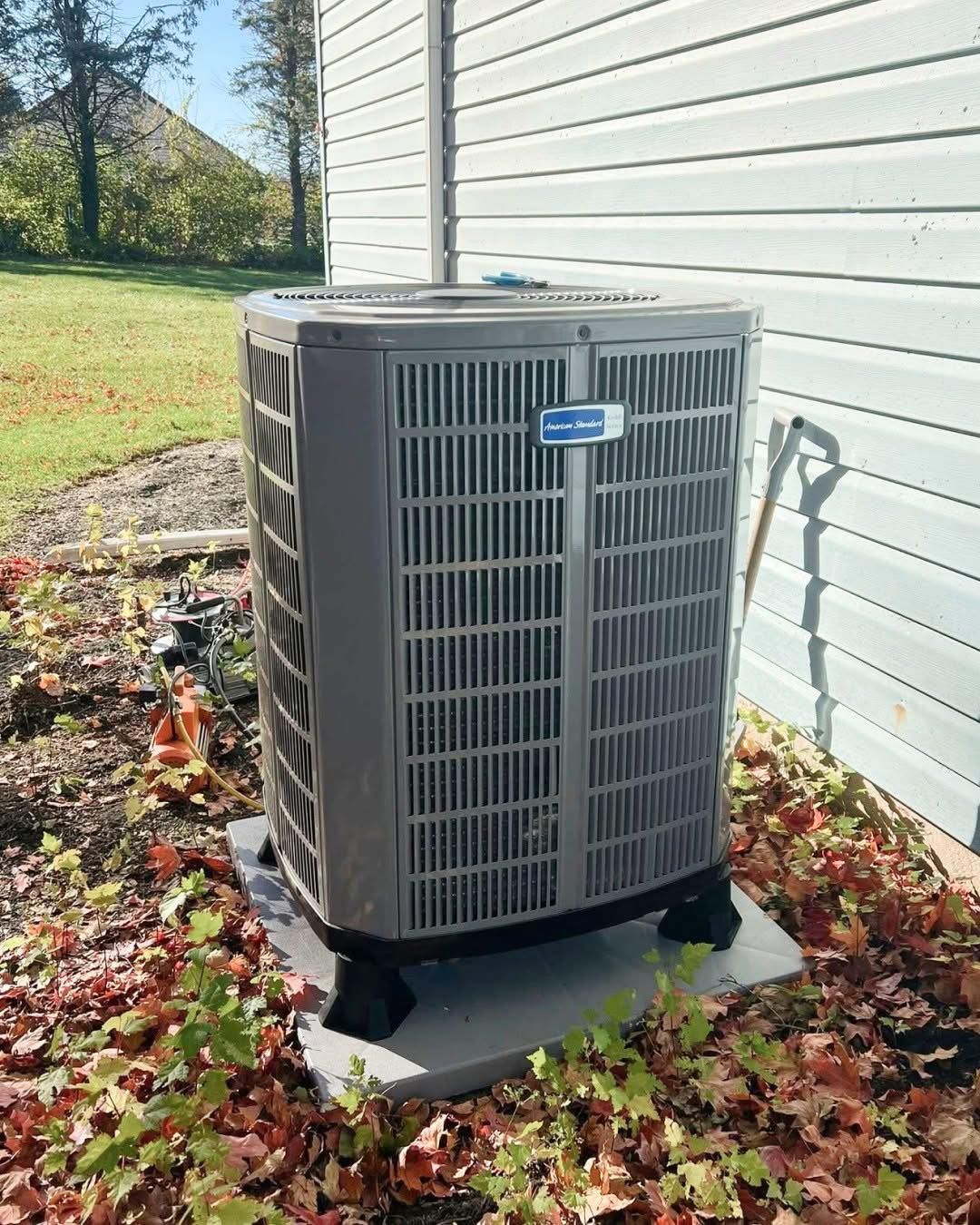 Outdoor air conditioner unit beside a white house wall, surrounded by fallen leaves on a concrete pad