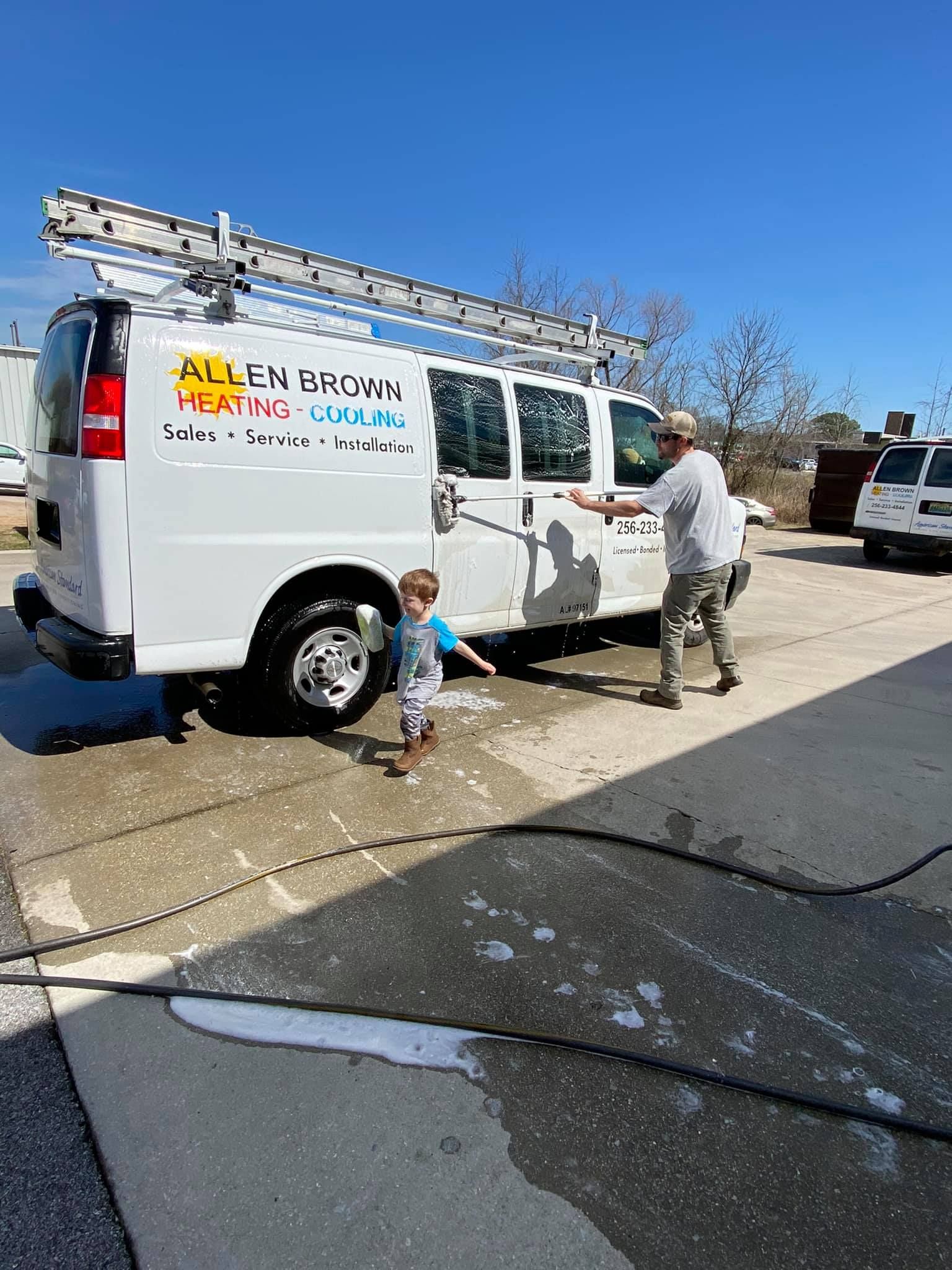 Worker spraying a white van with soapy water on a driveway, with a child nearby and a ladder on top.