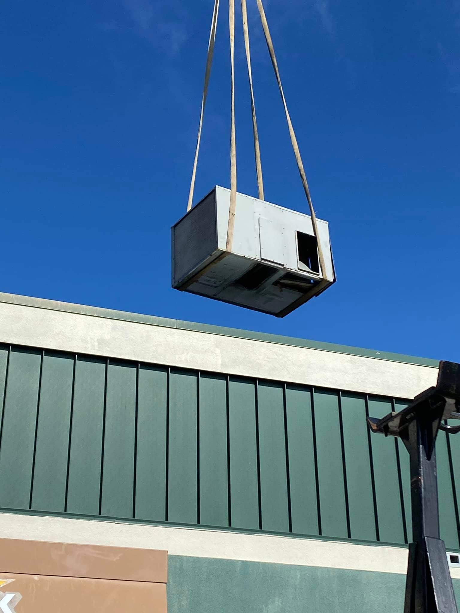 Crane lifting a white HVAC unit beside a green industrial building under a clear blue sky