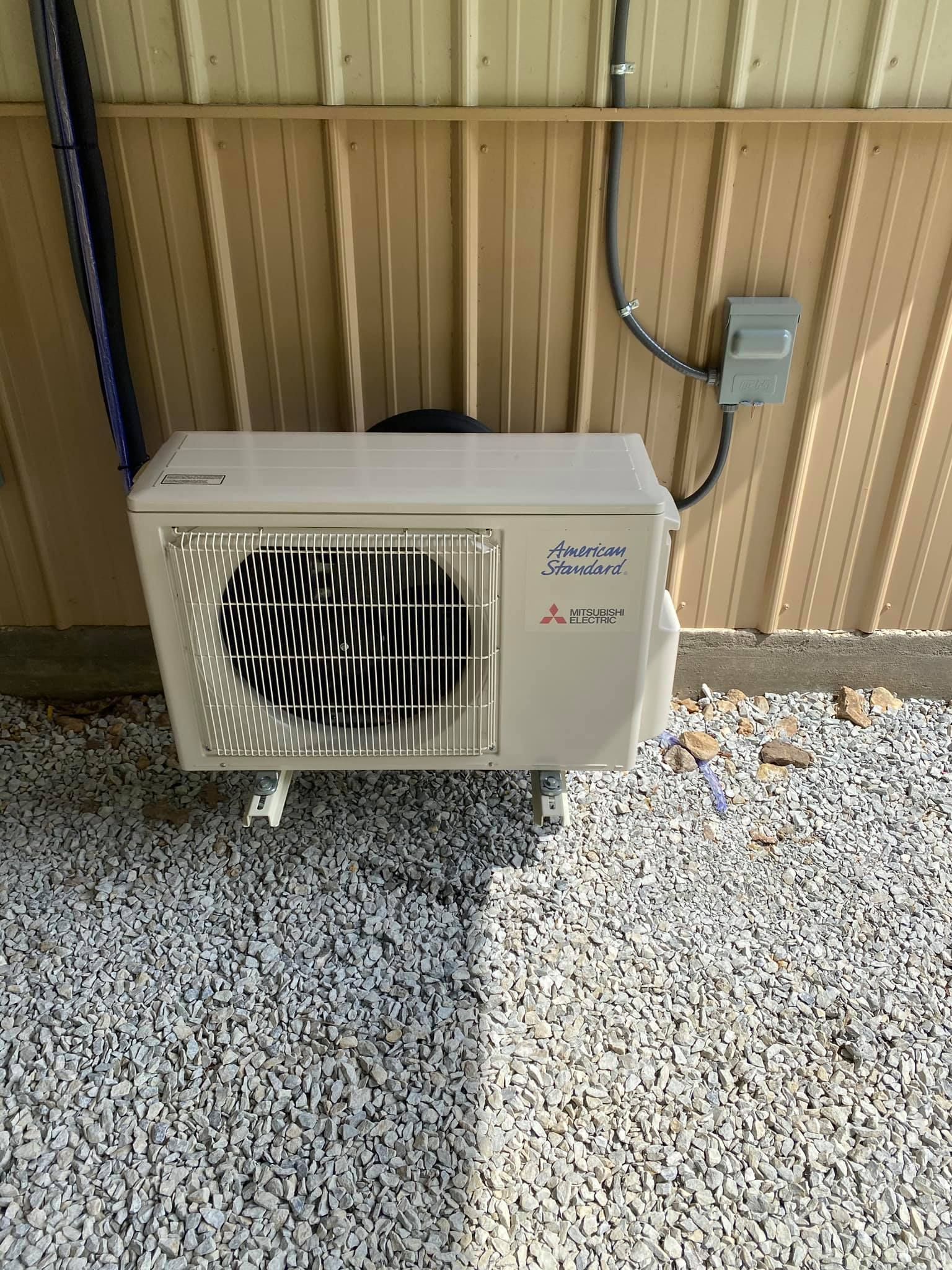 Outdoor air conditioner unit beside a tan wall, with gravel ground and a nearby electrical box