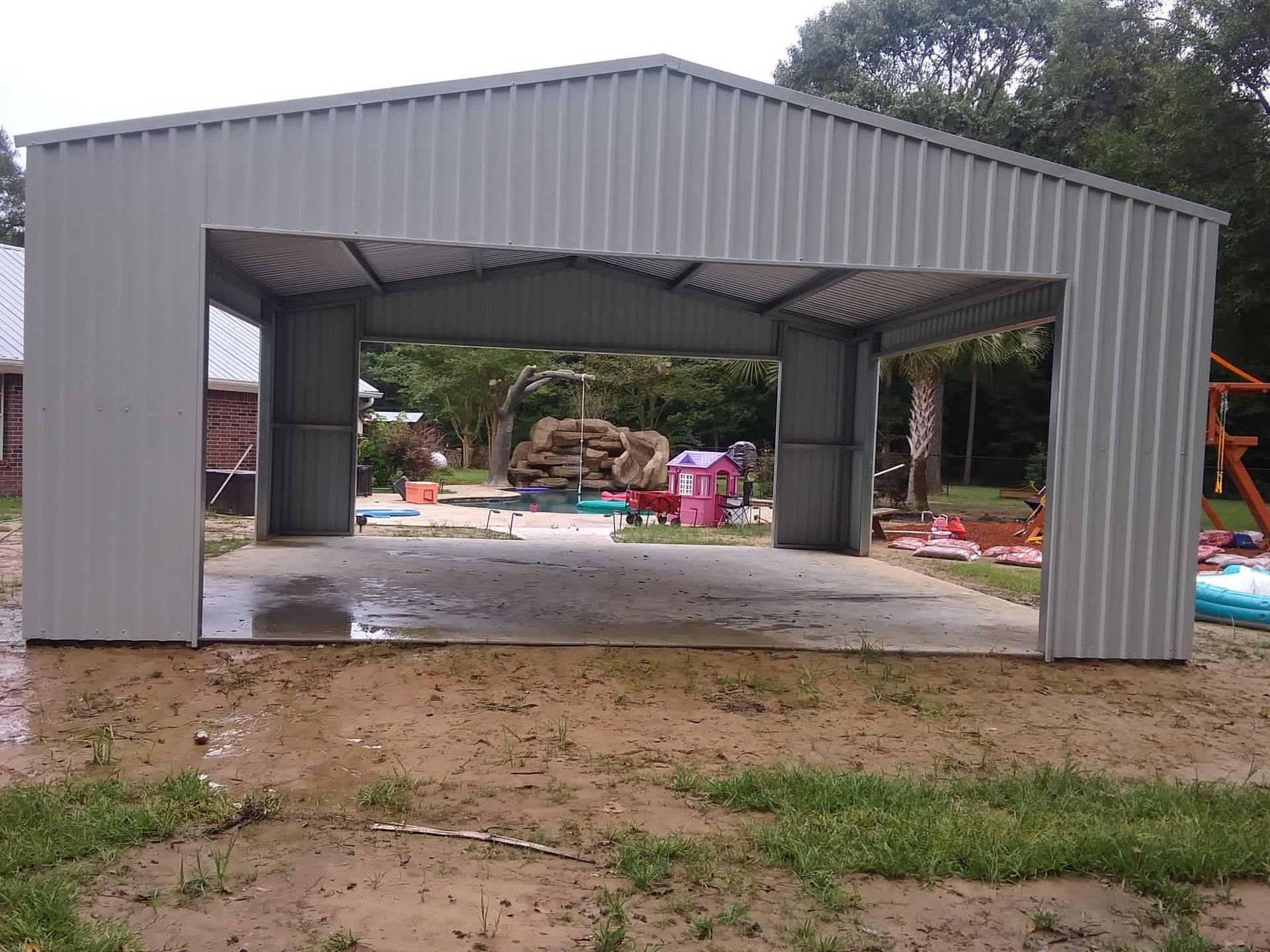 Gray metal open-sided shed with a concrete floor, outdoors on a dirt lot near trees and construction materials
