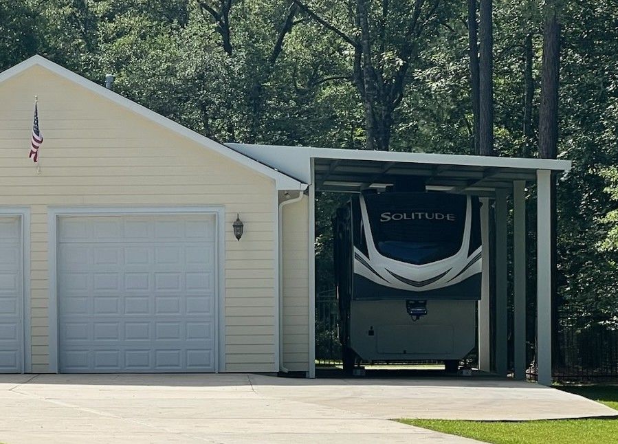 Garage with RV parked inside a carport next to a beige house, with an American flag at the front.