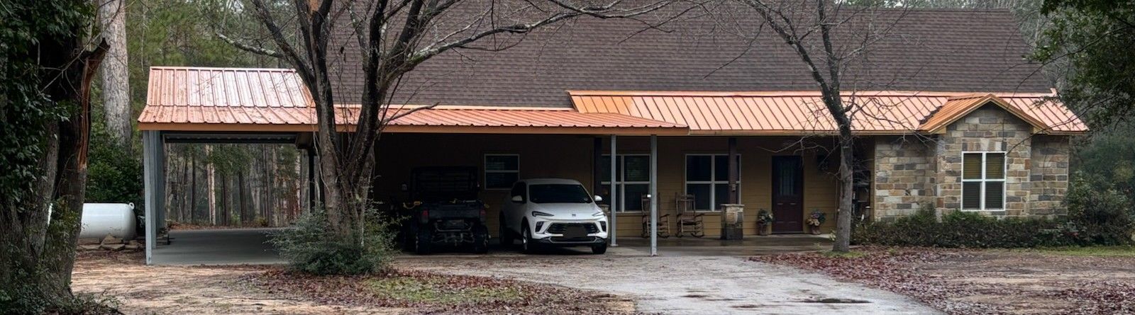 Single-story brick house with a carport and parked SUV under trees on a rainy day