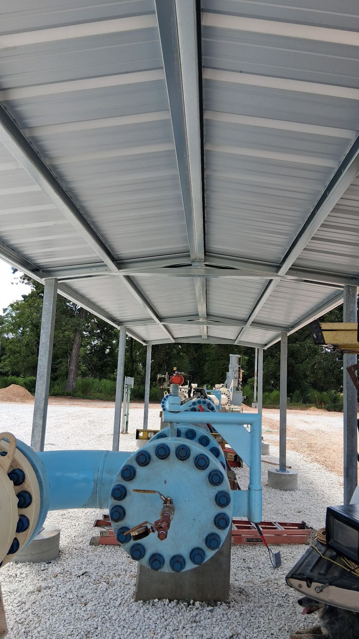 Under a metal roof, large blue industrial pipes and flanges sit on a concrete platform outdoors.