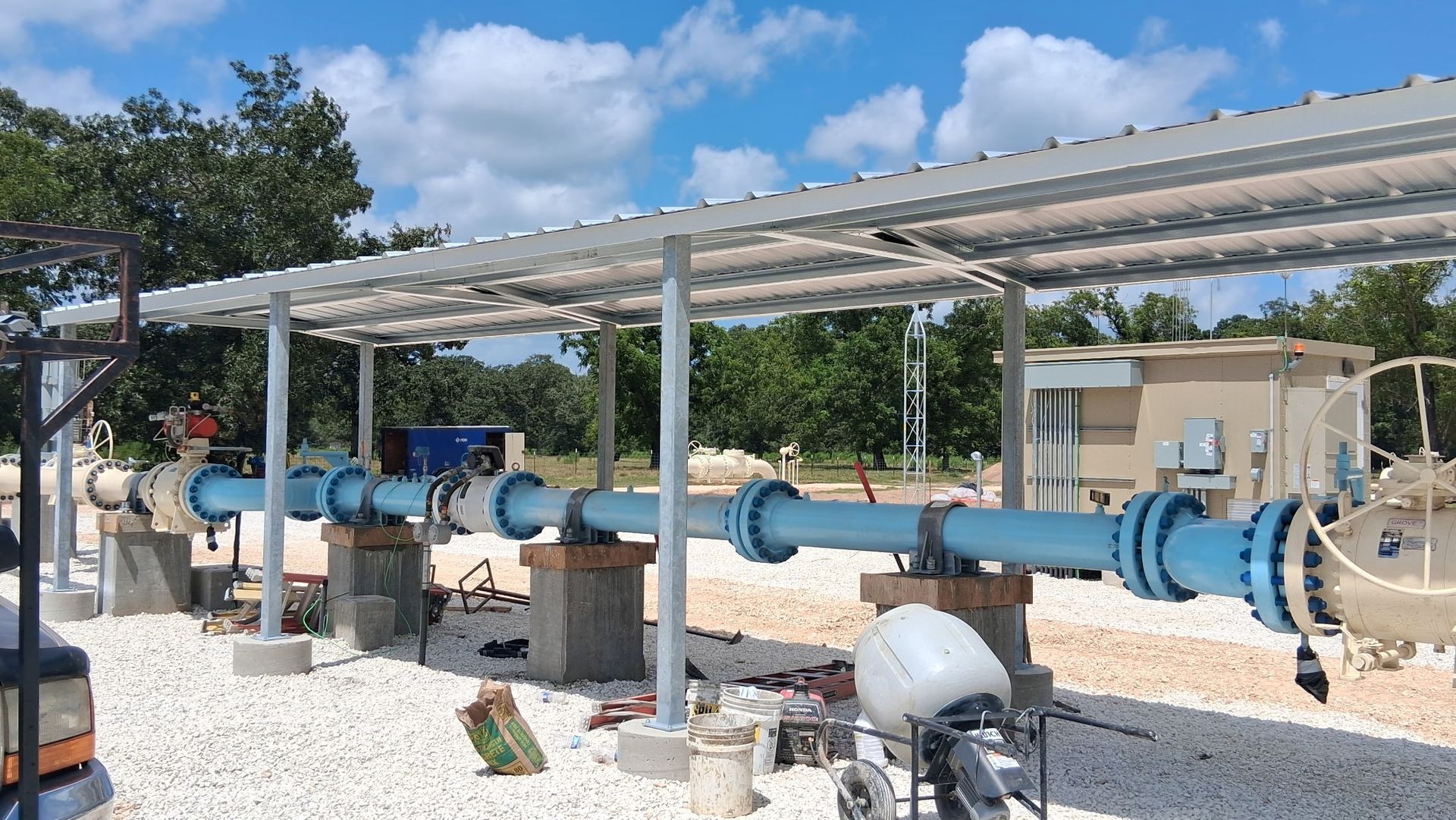 Blue industrial pipes and valves under a metal shelter on a gravel utility site under a sunny sky