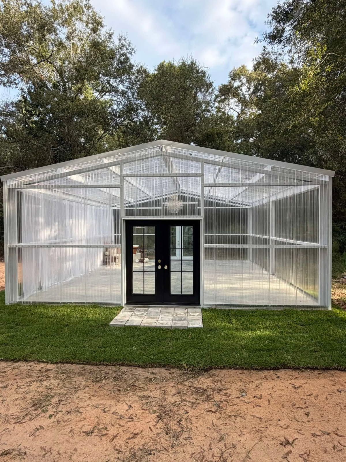 Small glass greenhouse with a black double door, set on grass with trees in the background.