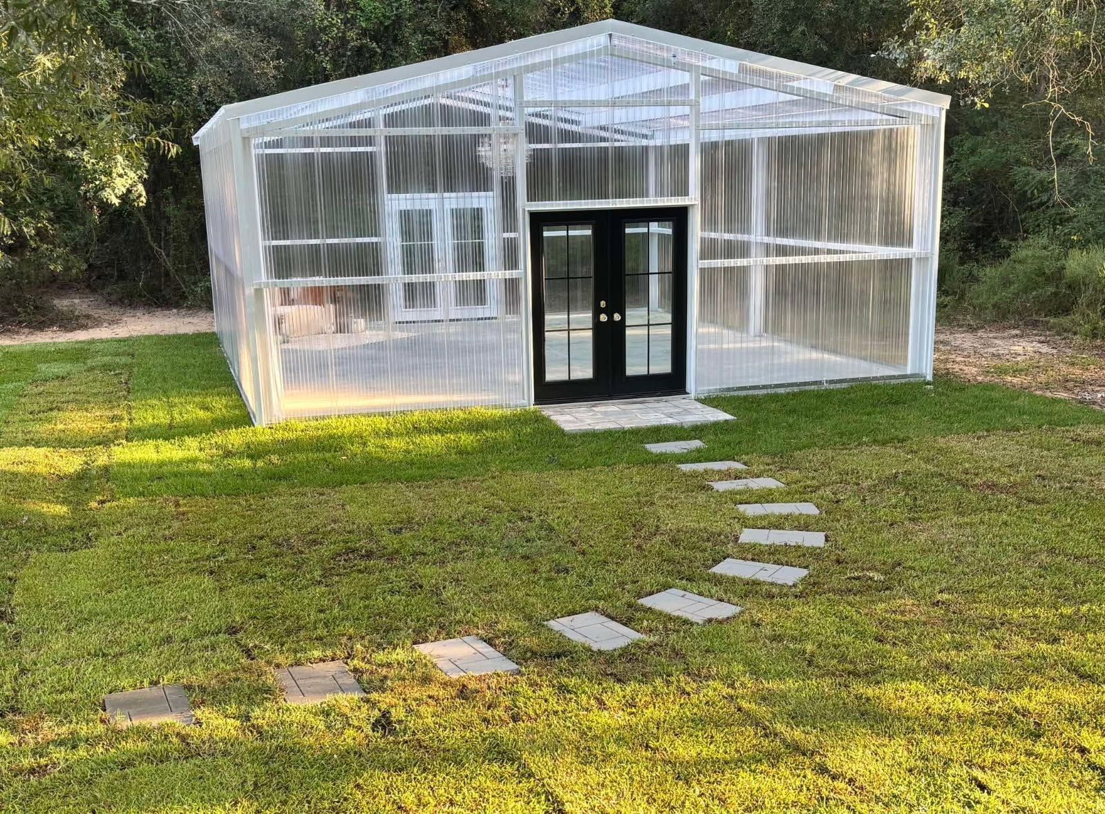 Small transparent greenhouse with black double doors on a grassy yard, with stepping stones leading to it