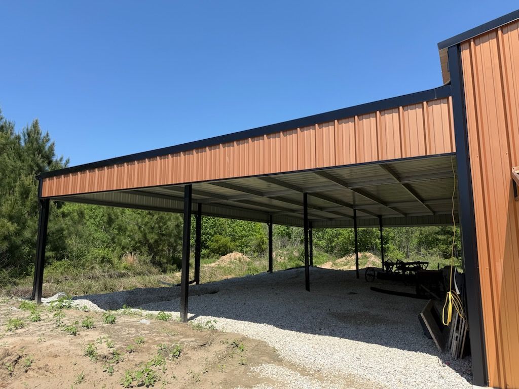 Brown metal carport with black supports on gravel ground, in front of trees.