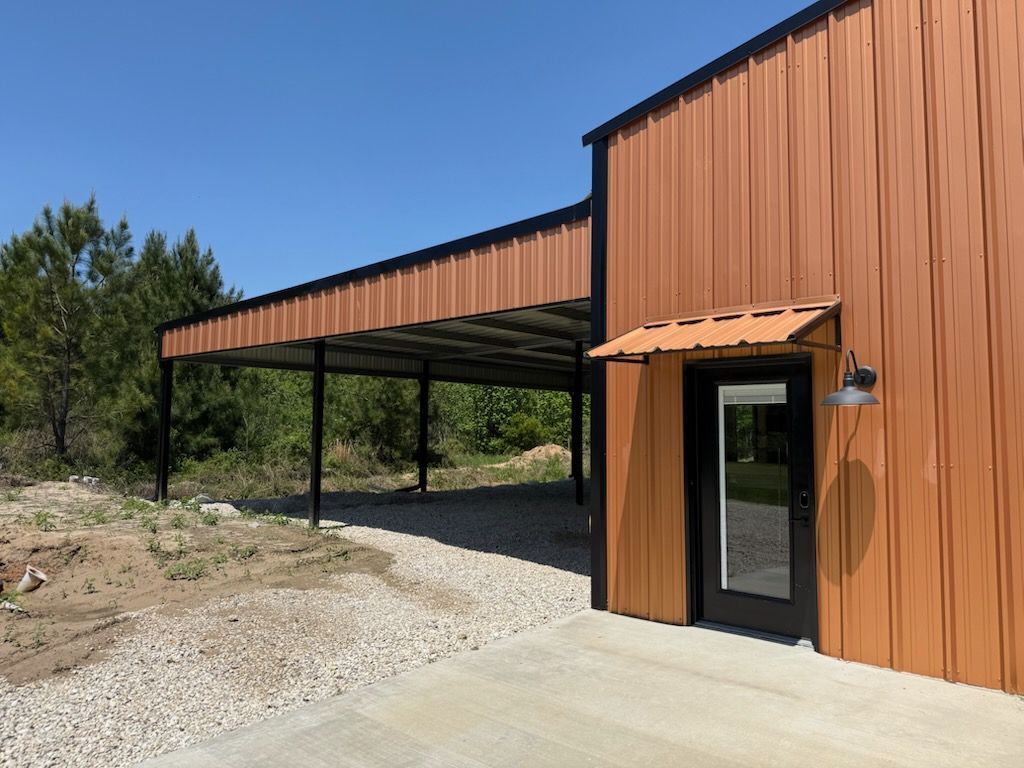Brown metal-clad building with a covered carport. Gravel driveway and a concrete patio.