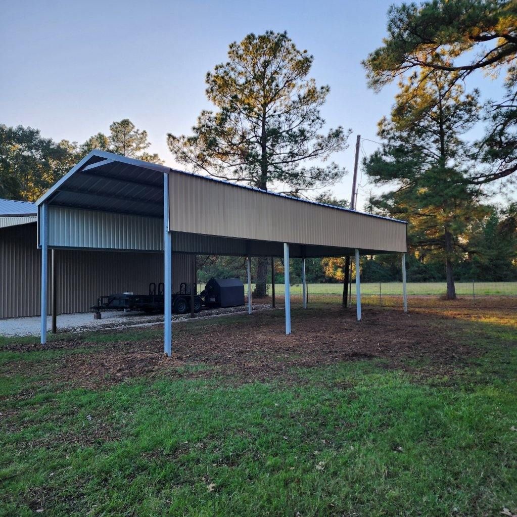 Metal carport with parked equipment, set in a grassy yard, trees in the background under a blue sky.