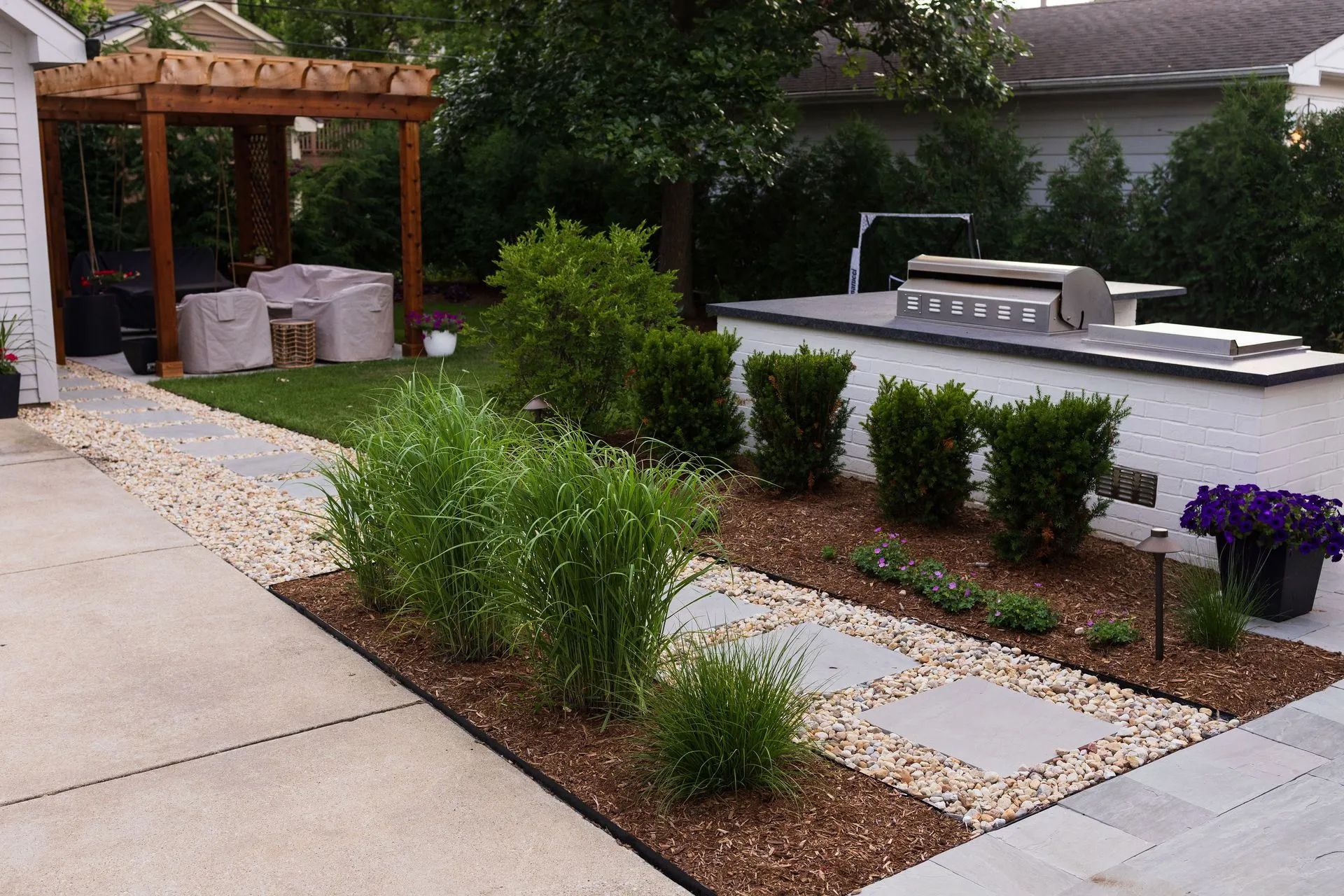 Backyard patio with gravel path, plants, grill station, and wooden pergola.
