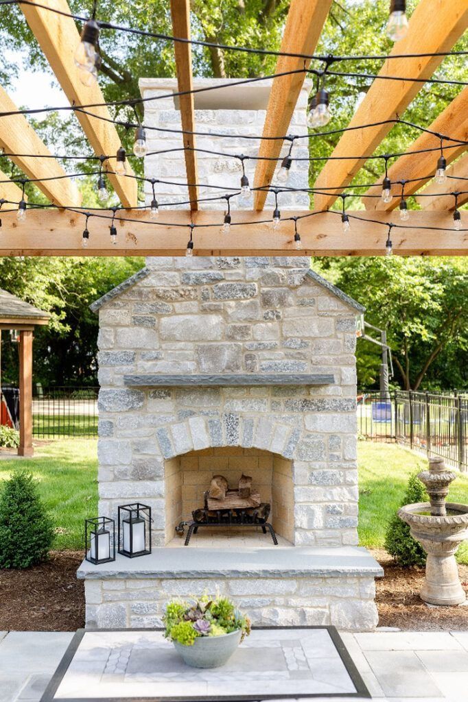 Stone outdoor fireplace with pergola overhead, lights, and greenery in a backyard setting.