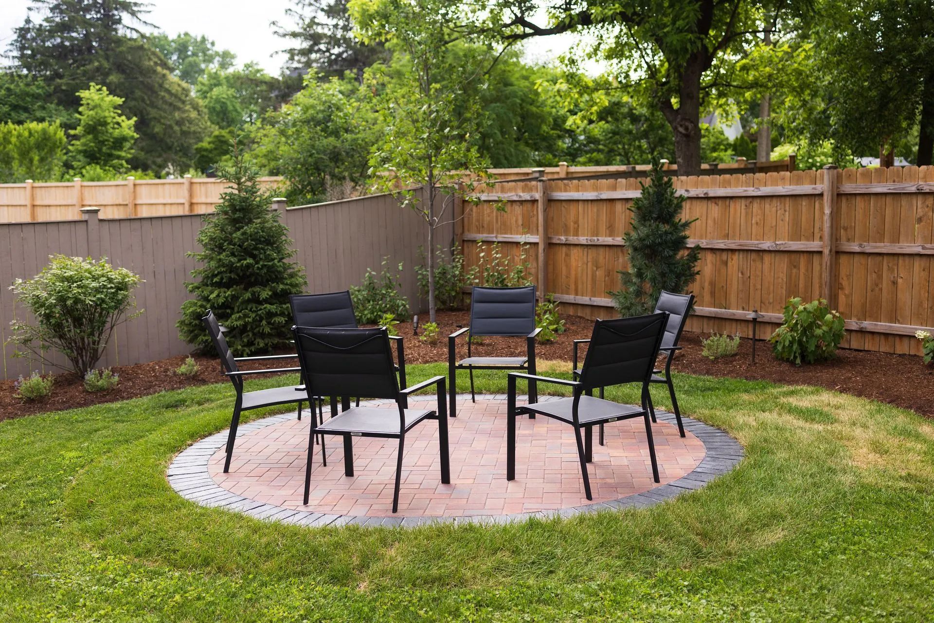 A backyard patio with chairs arranged around a circular brick area surrounded by grass.