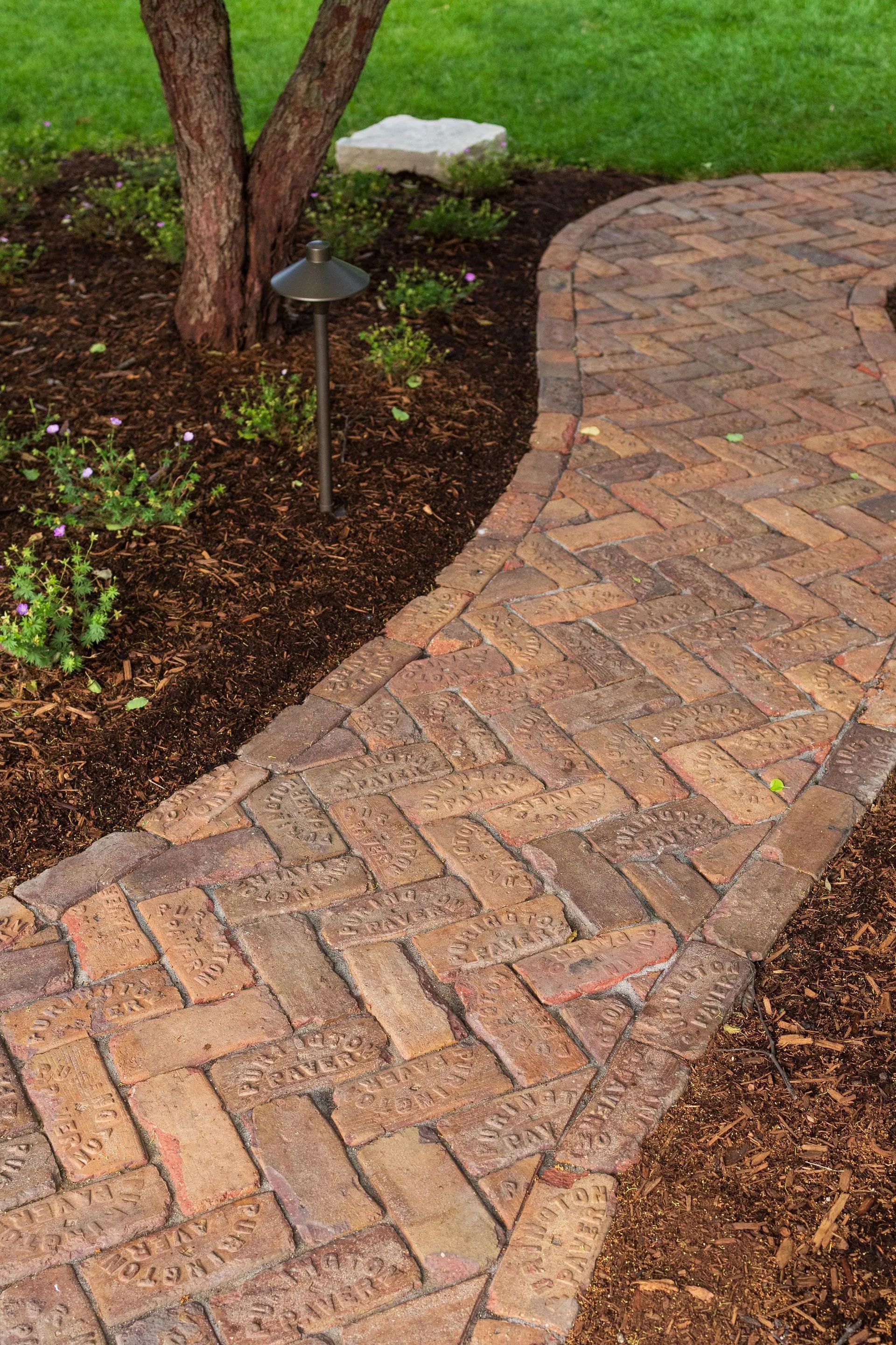 Brick pathway curves through a garden bed with a small tree and green lawn.