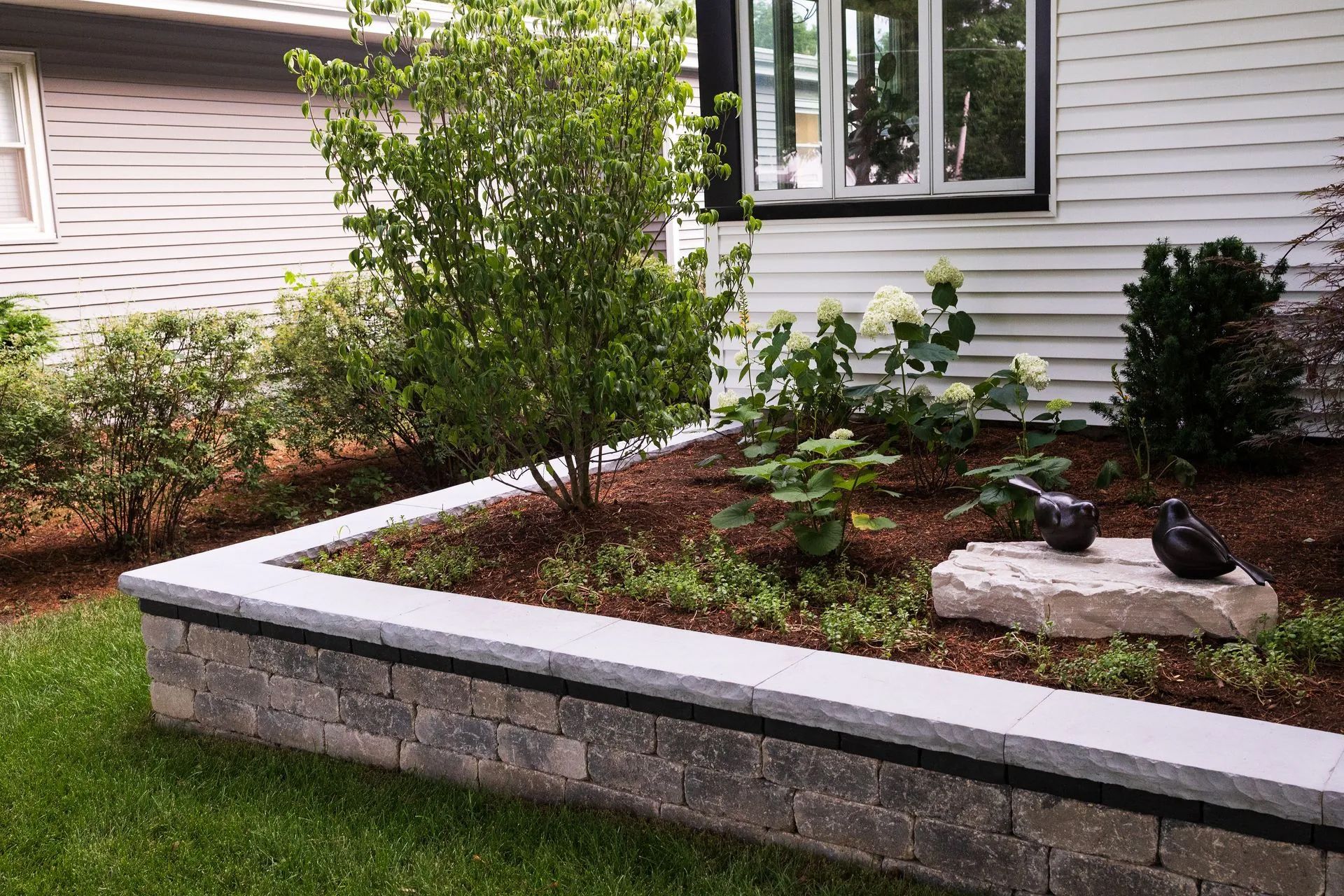 Stone-retained garden bed with various green plants and mulch, beside a house with white siding and a black window frame.