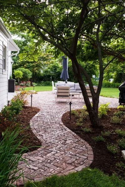 Brick path curves through a garden to a patio with outdoor seating. Tree shades the path.