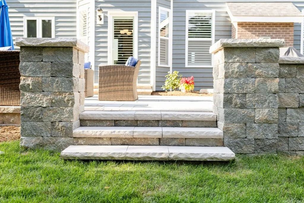 Stone steps lead to a patio with a house in the background, surrounded by green grass.