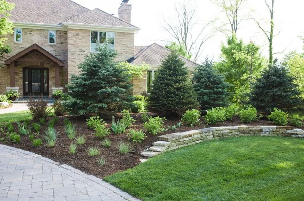 Brick house with landscaped front yard, featuring trees, shrubs, and a stone retaining wall.