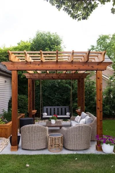 Pergola with swing and wicker seating on a patio, surrounded by greenery and flowers.
