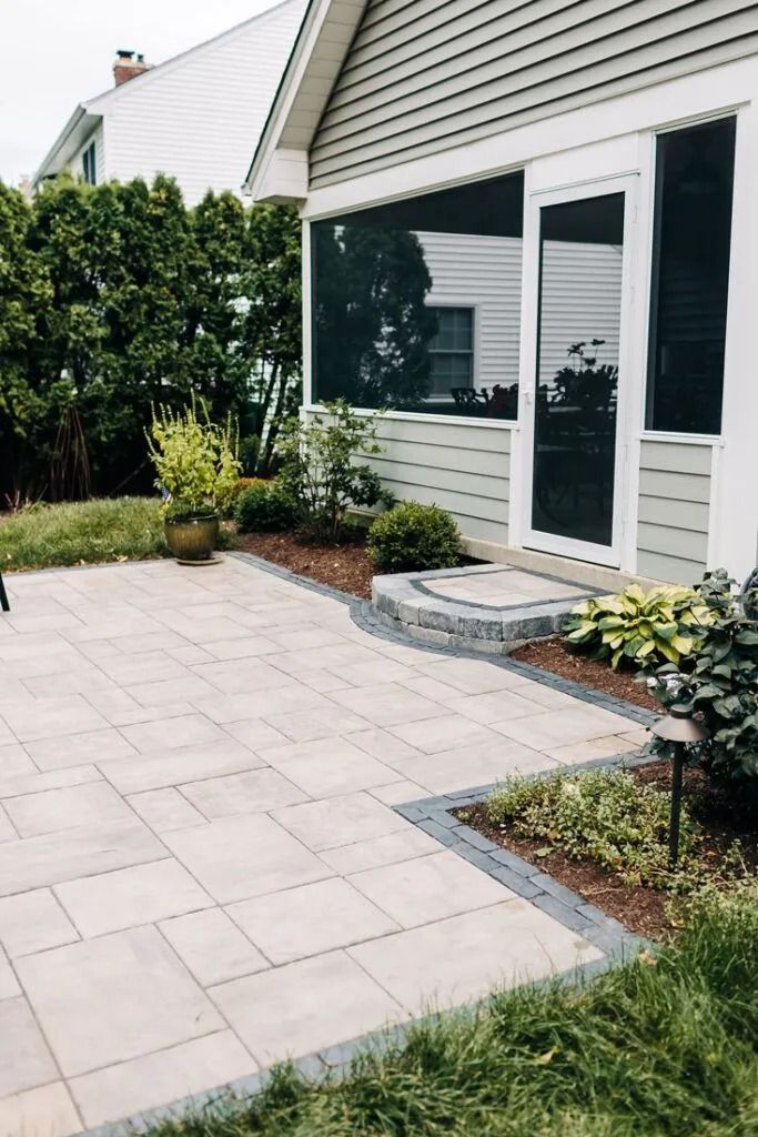 Stone patio leading to a screened porch with landscaping. Beige pavers edged in dark gray.