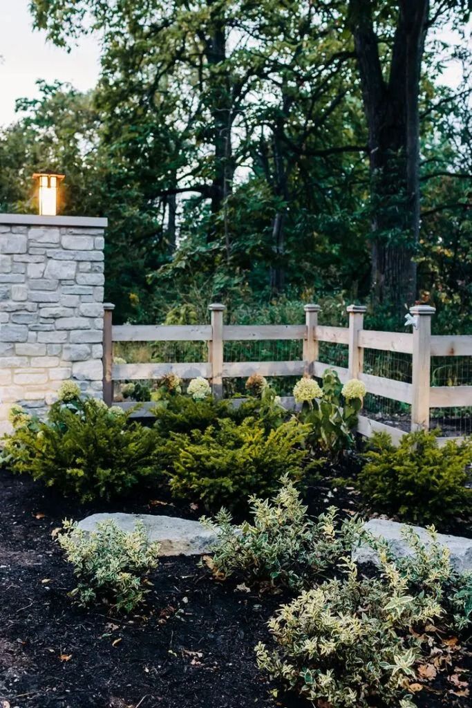 Stone pillar with light, split-rail fence, garden with green bushes, dark mulch, and trees in background.