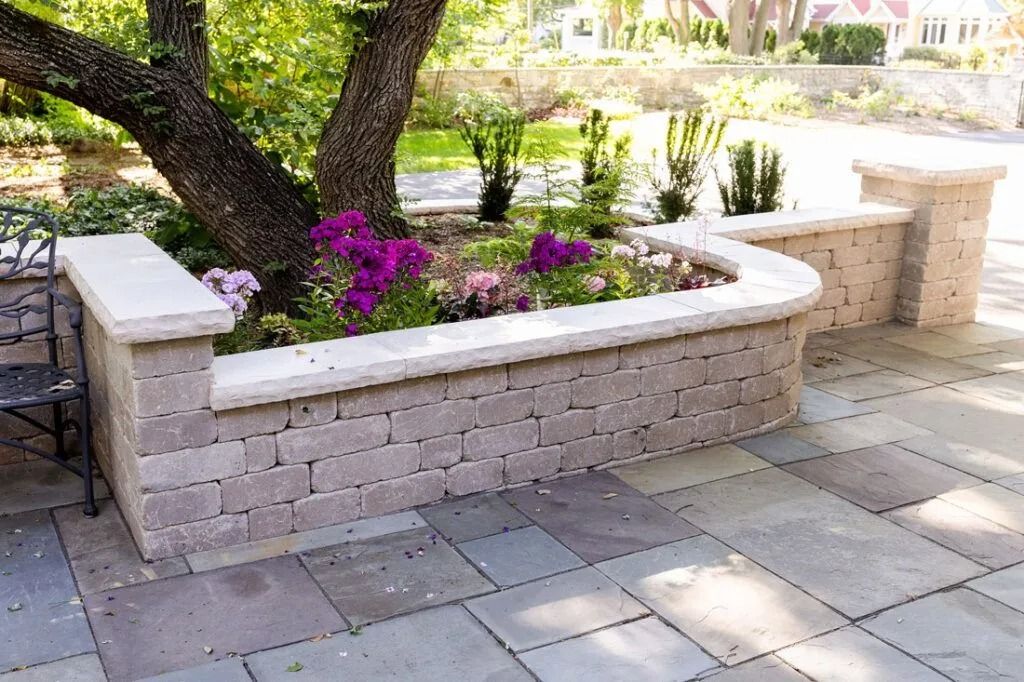 Stone retaining wall with tree and flowers in a paved patio.