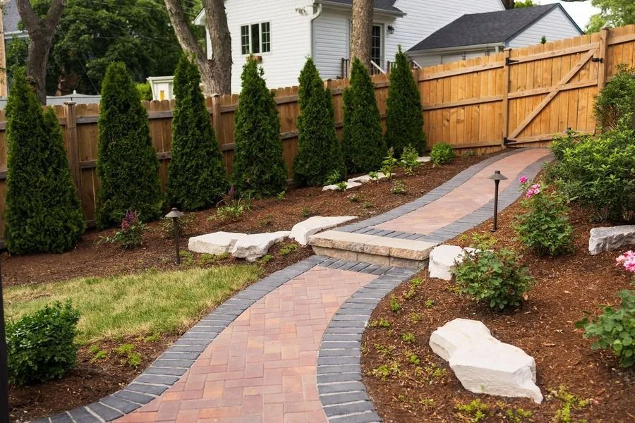 Brick pathway ascends a sloped yard lined with evergreens and a wooden fence; stone accents.