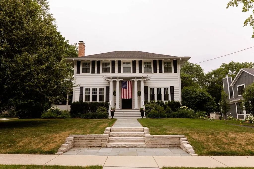 White two-story house with American flag. Green lawn, stone steps, black shutters. Cloudy day.