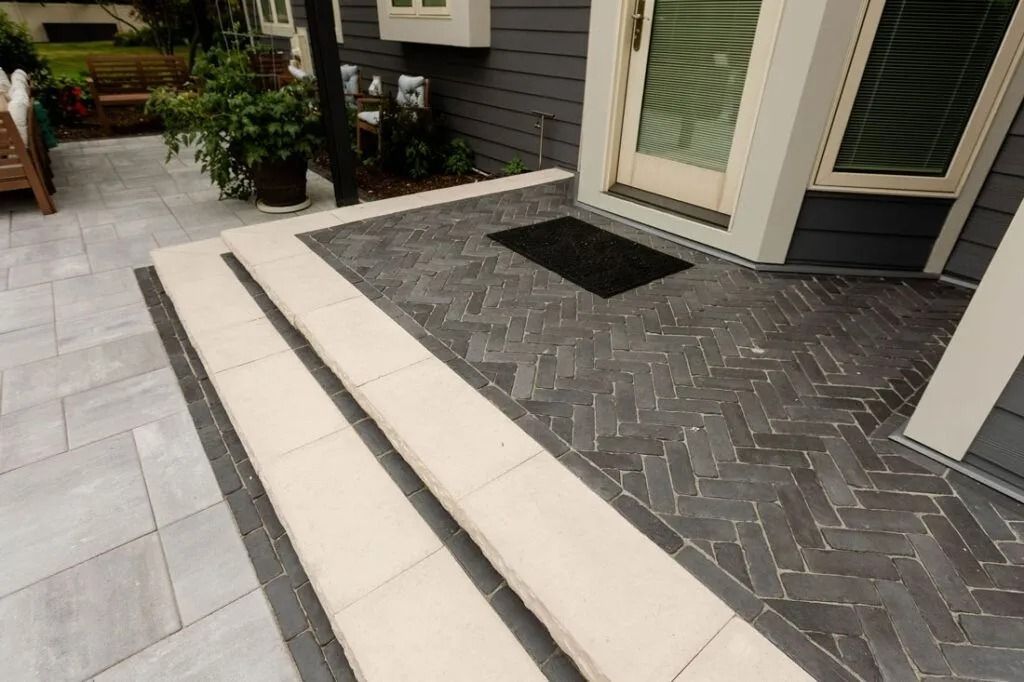Steps leading to a home with herringbone brick patio, light-colored step facing, and dark siding.