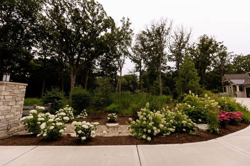 Landscaped yard with white and pink flowers, trees, and a stone entrance post.