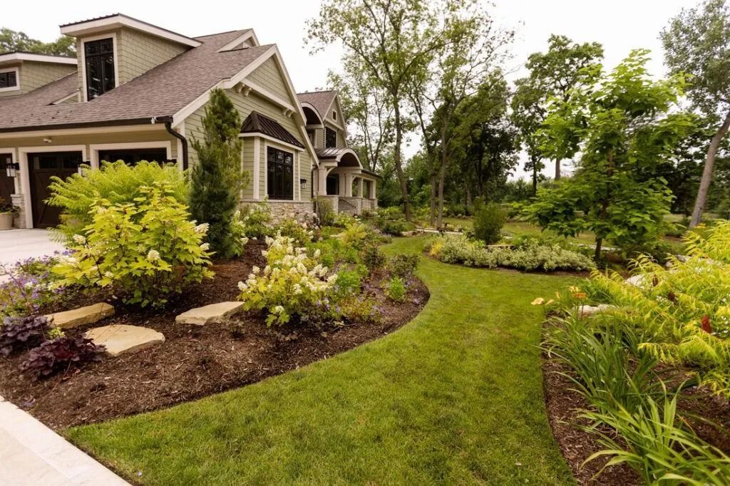 A winding green grass path leads through a lush garden toward a two-story house with green siding and a dark roof.
