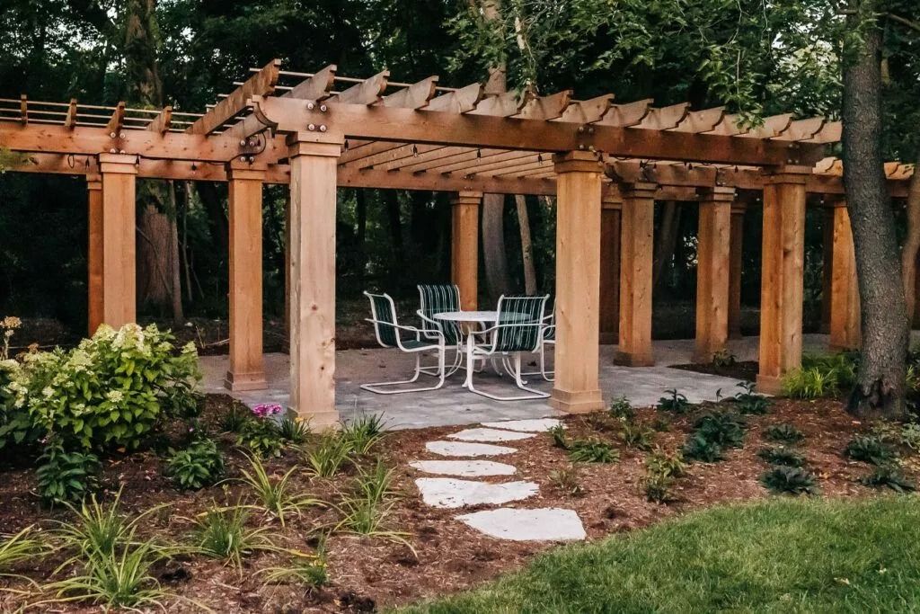 Pergola with dining table and chairs in a garden setting, stone path, trees and bushes.