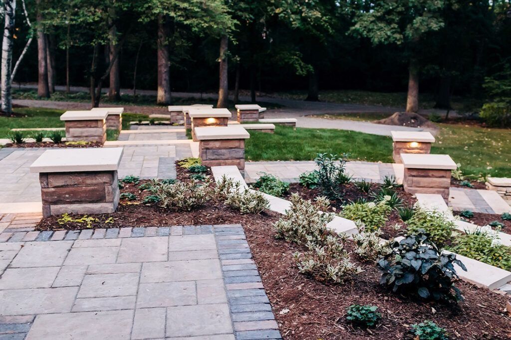 Brick and stone patio with tiered planters, lit pillars, and trees in background.