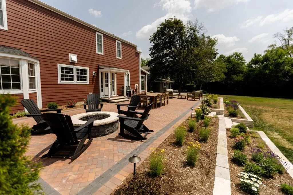 Brick patio with fire pit, Adirondack chairs, and dining table near red brick house and a grassy lawn.