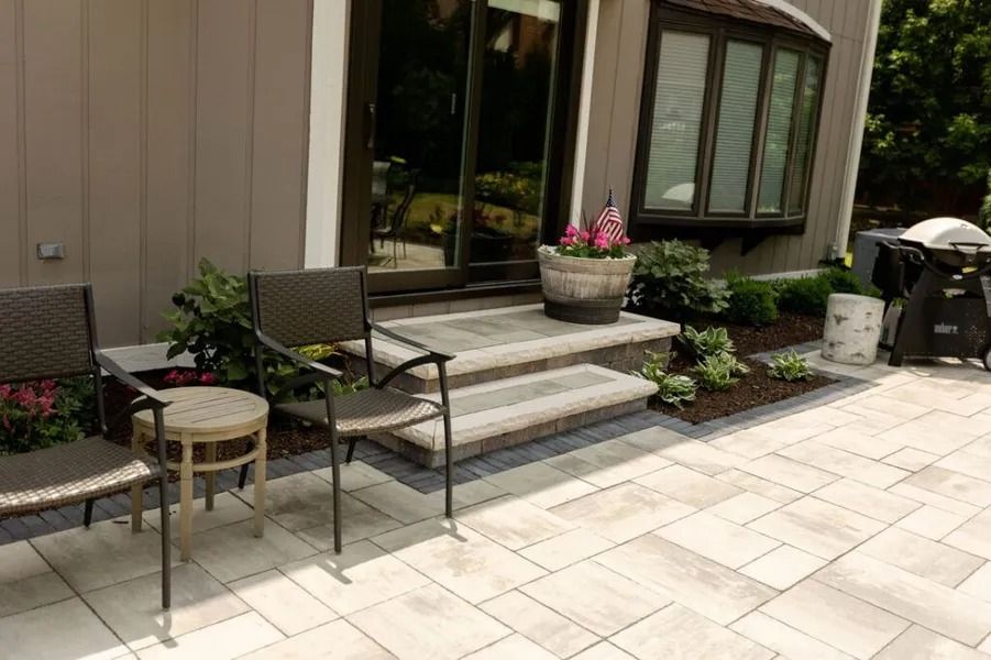 Patio with gray pavers, steps, and chairs. Doorway leads to the house, with potted flowers.