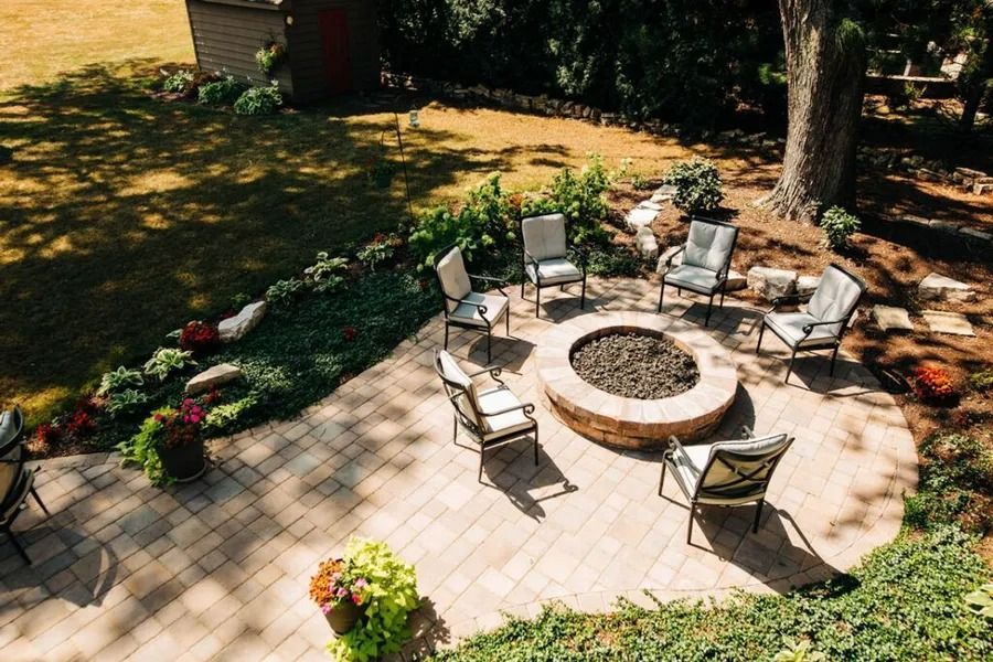 Backyard patio with fire pit surrounded by chairs, greenery, and trees.