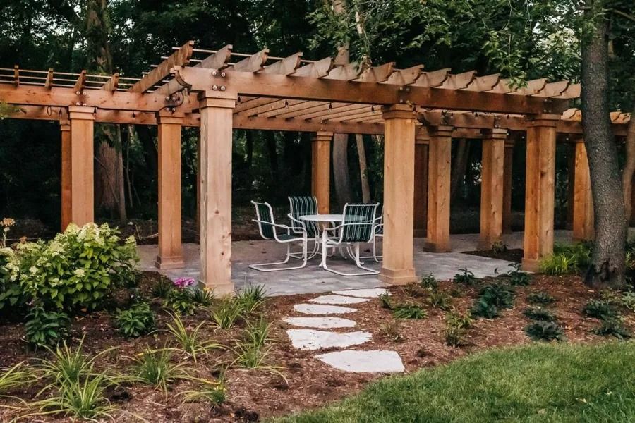 Wooden pergola with a table and chairs on a stone patio, path leading from a grassy area.