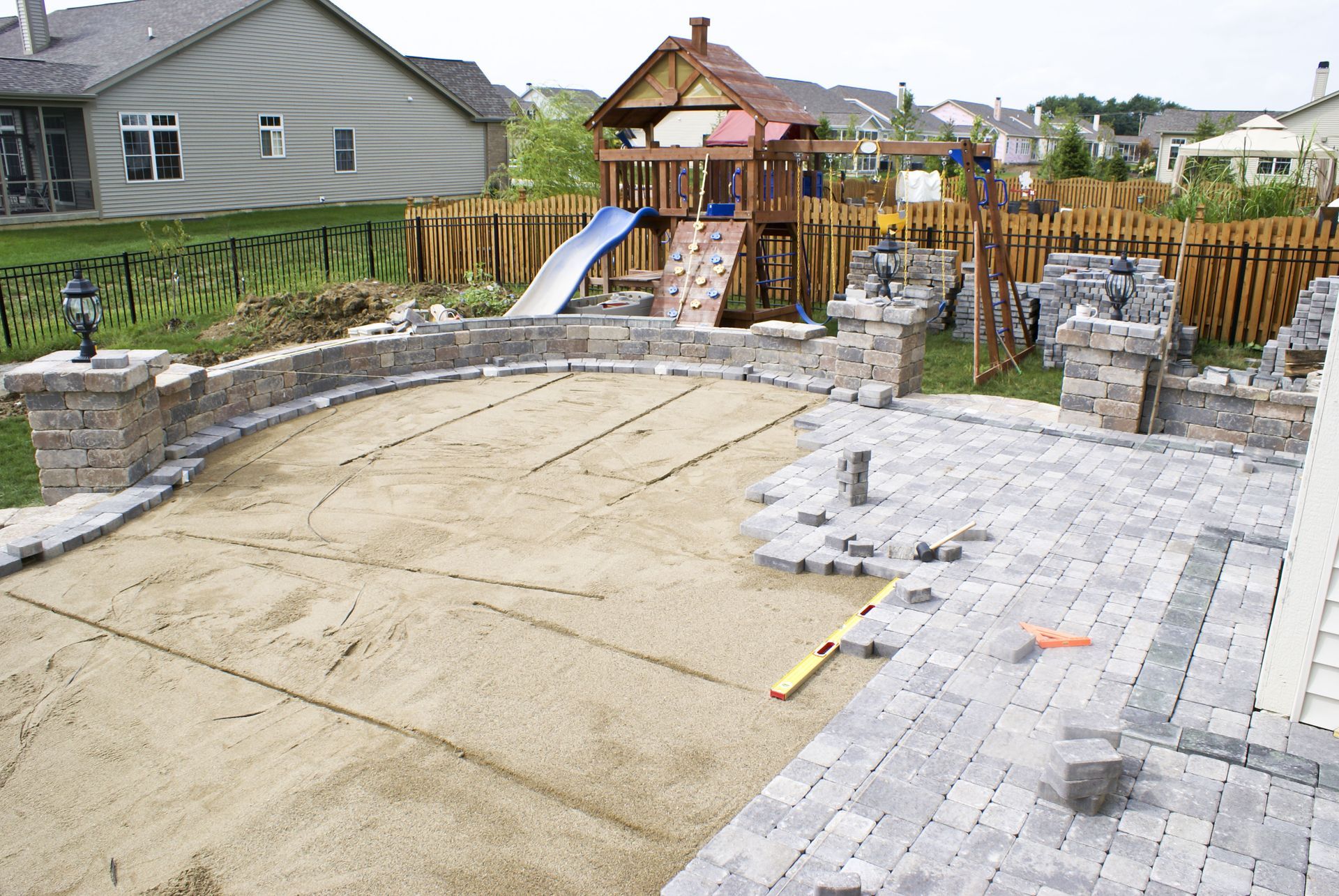 Backyard patio under construction; sand base, stone borders, playground, stone walls, and pavers being laid.